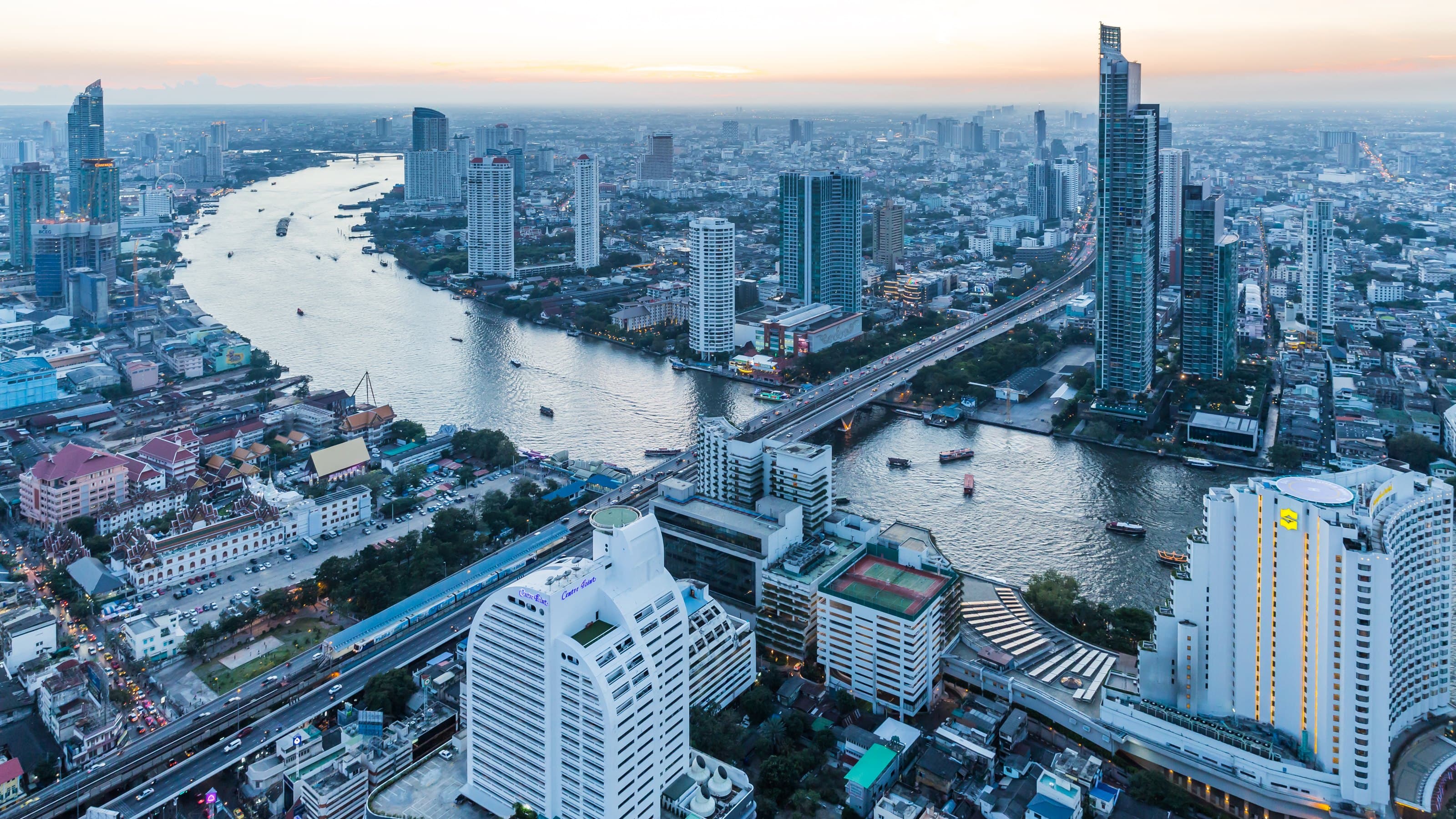 The Chao Phraya river curving through Bangkok's Bang Rak business district at dusk, office towers and an elevated Skytrain line lit along the riverbank, Bangkok, Thailand
