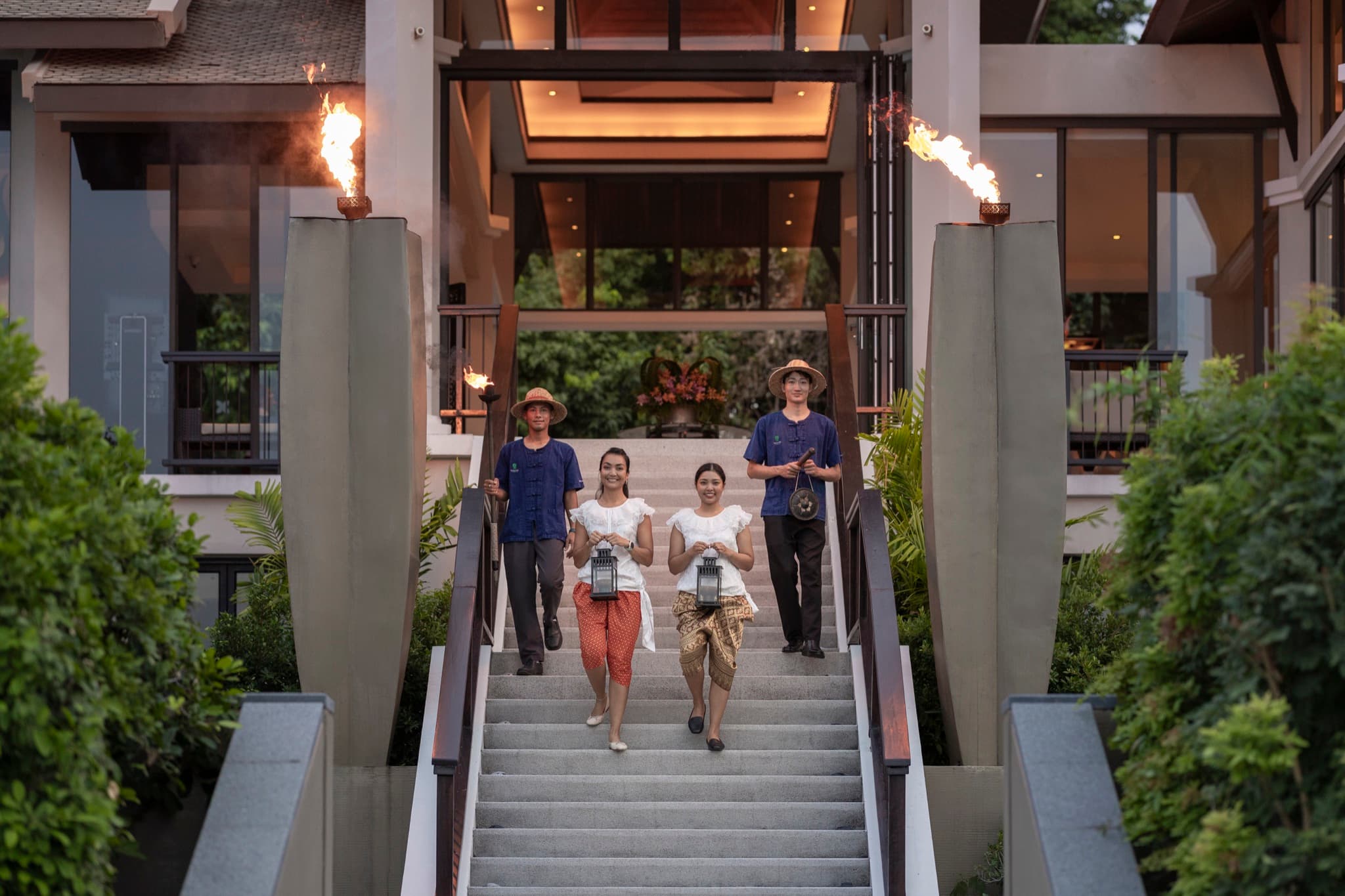 Banyan Tree Samui staff in traditional Thai dress descending a torch-lit staircase to welcome arriving guests at dusk, Koh Samui, Thailand