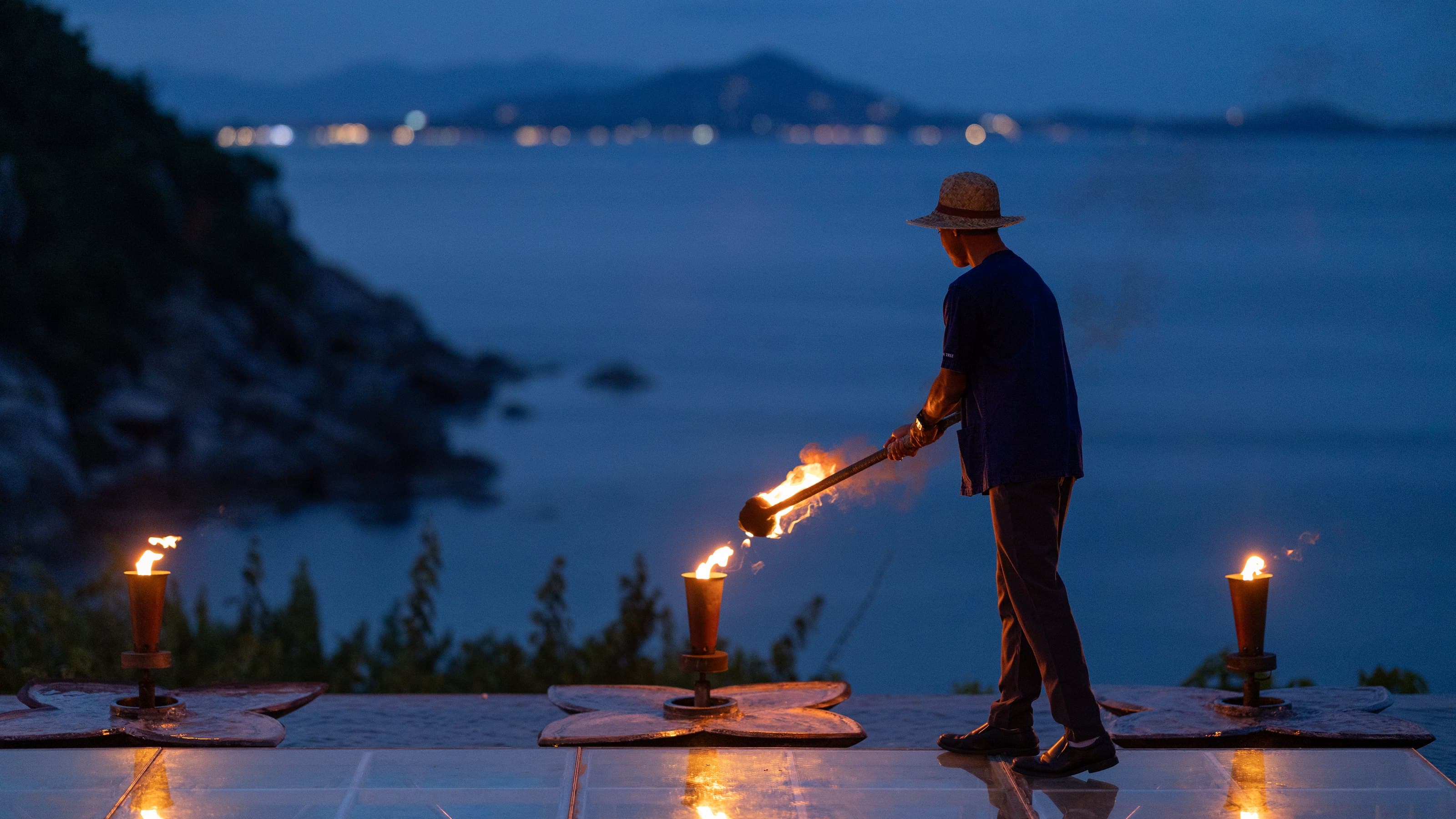 A Banyan Tree Samui host lighting the torch line along the water's edge at dusk, Koh Samui, Thailand