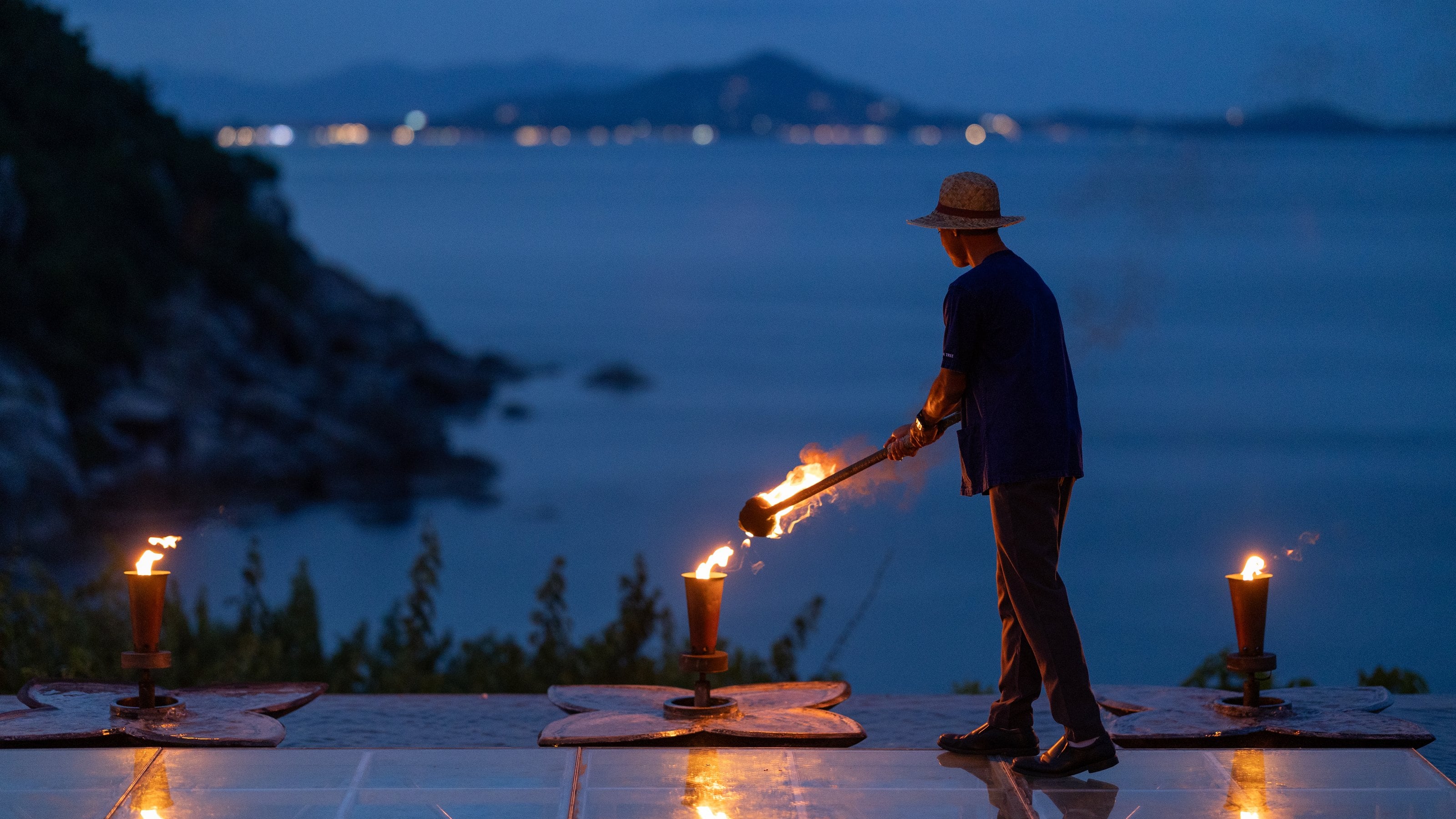 A Banyan Tree Samui host lighting the torch line along the water's edge at dusk, Koh Samui, Thailand