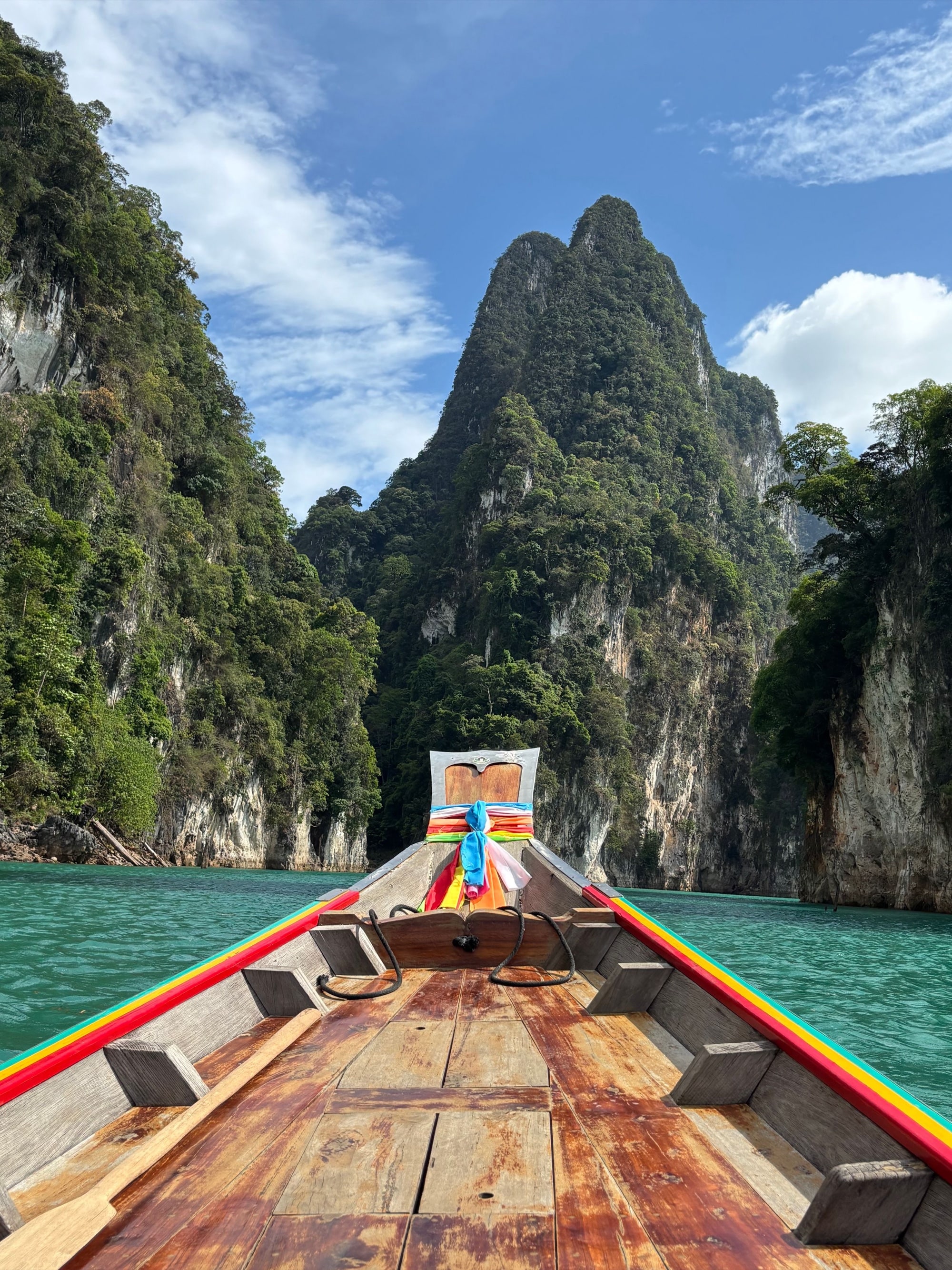 The view from the bow of a traditional Thai longtail boat crossing an emerald lagoon toward towering jungle-clad limestone karst peaks, Phang Nga, Thailand